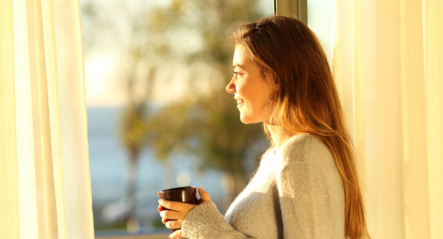 A woman holding a mug looking out the window during golden hour
