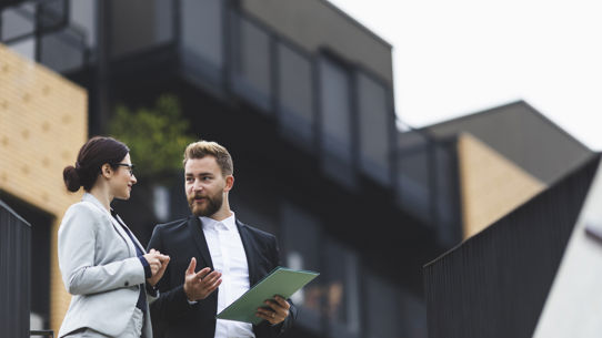 Two people in suits talking in front of an apartment building